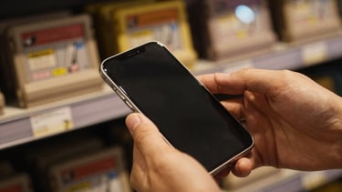 Moody close-up of hands inspecting a certified pre-owned mobile screen inside a game shop, warm gold lighting, retro game cartridges blurred in the background